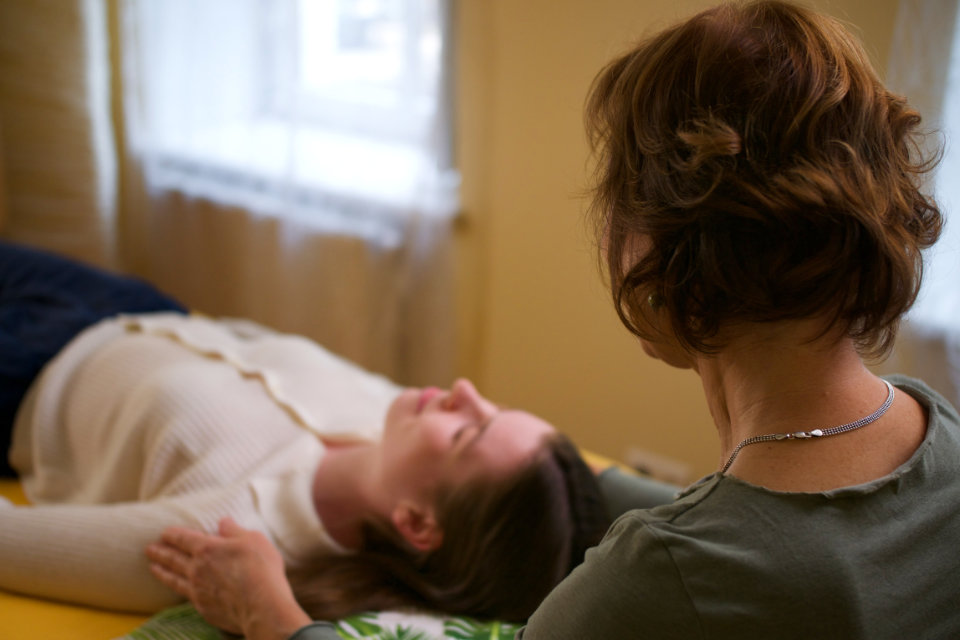 Nahaufnahme einer craniosacralen Behandlung: Klientin liegt entspannt auf der Liege, Praktikerin im Hintergrund